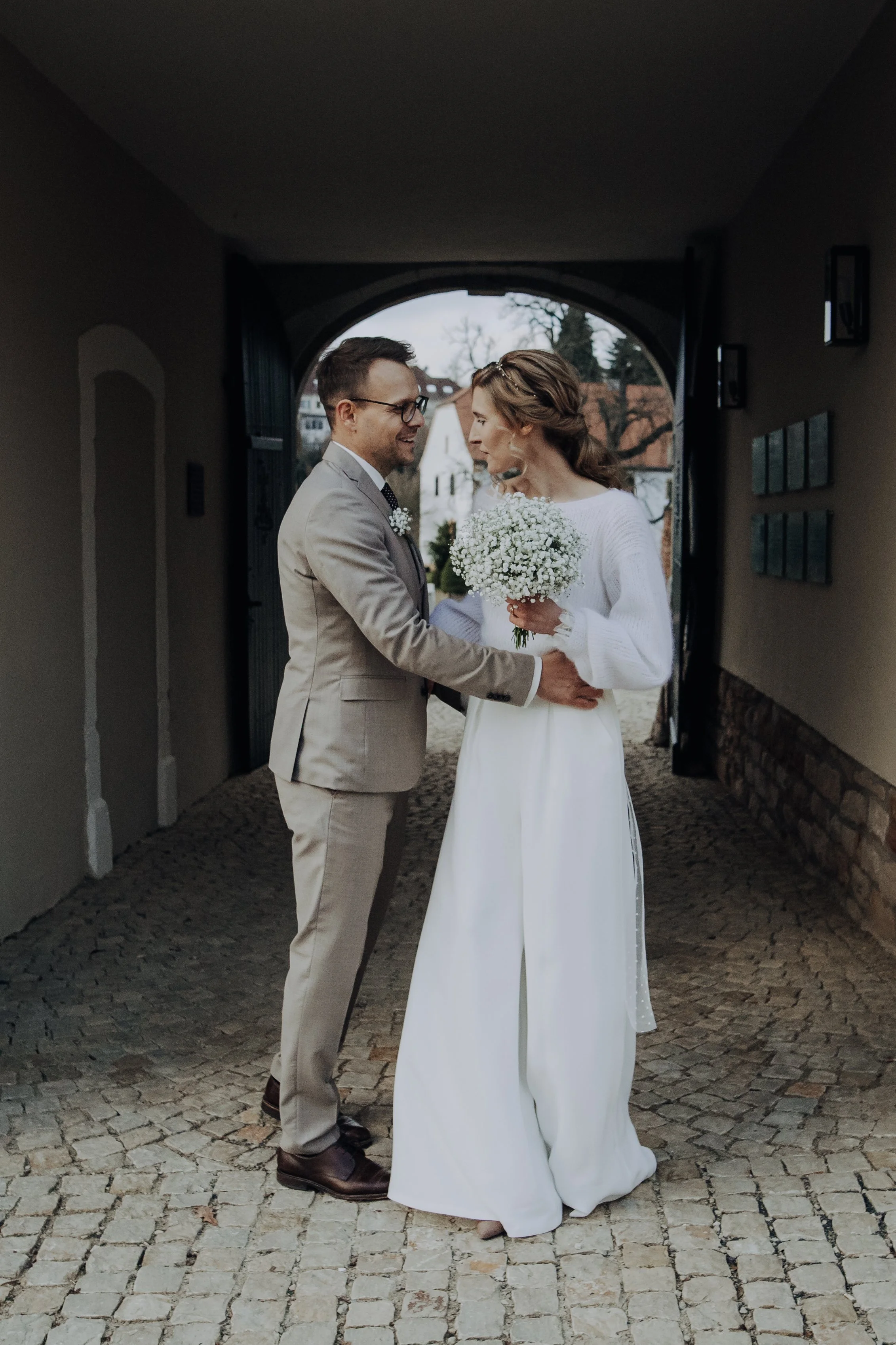Ein Brautpaar steht sich in einer unter einem Bogen gelegenen Passage gegenüber, lacht und hält sich an den Händen. Hochzeitsreportage Saarland, Brautpaarshooting Saarland, Hochzeitsfotografin Saarland, Fotografin Saarland.