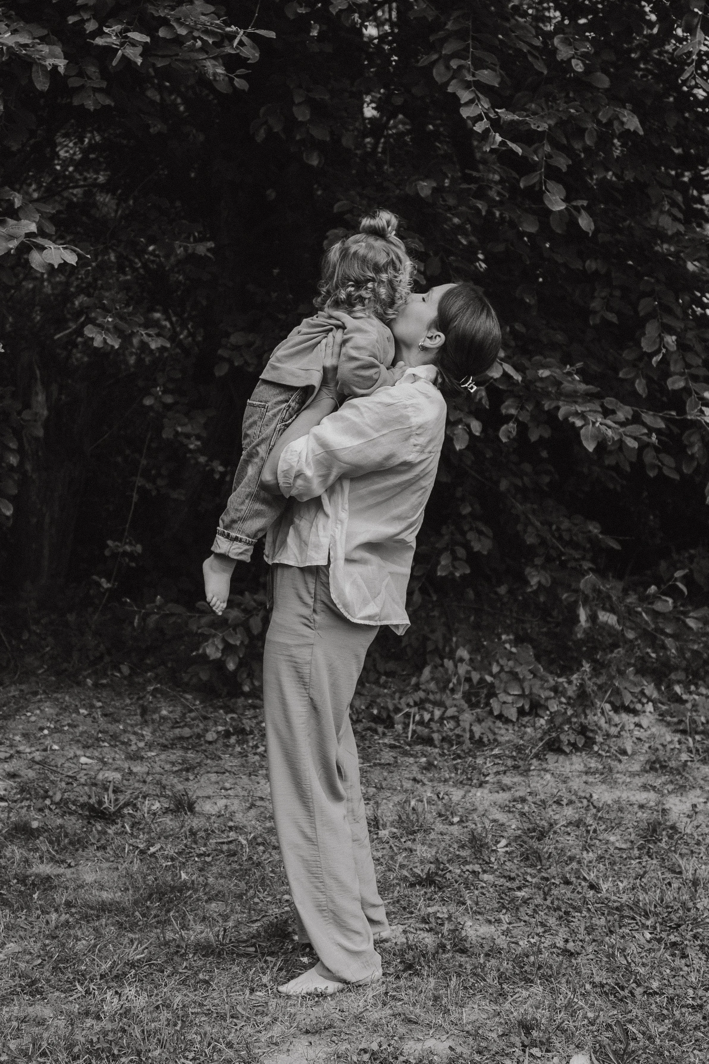 Eine Frau hebt ein Kind hoch und küsst es im Freien, umgeben von Bäumen, in einer schwarzen und weißen Fotografie. Mutter & Kind Shooting im Saarland, Familienshooting im Saarland, Familienfotografin. Fotografie für Mütter und Kinder.