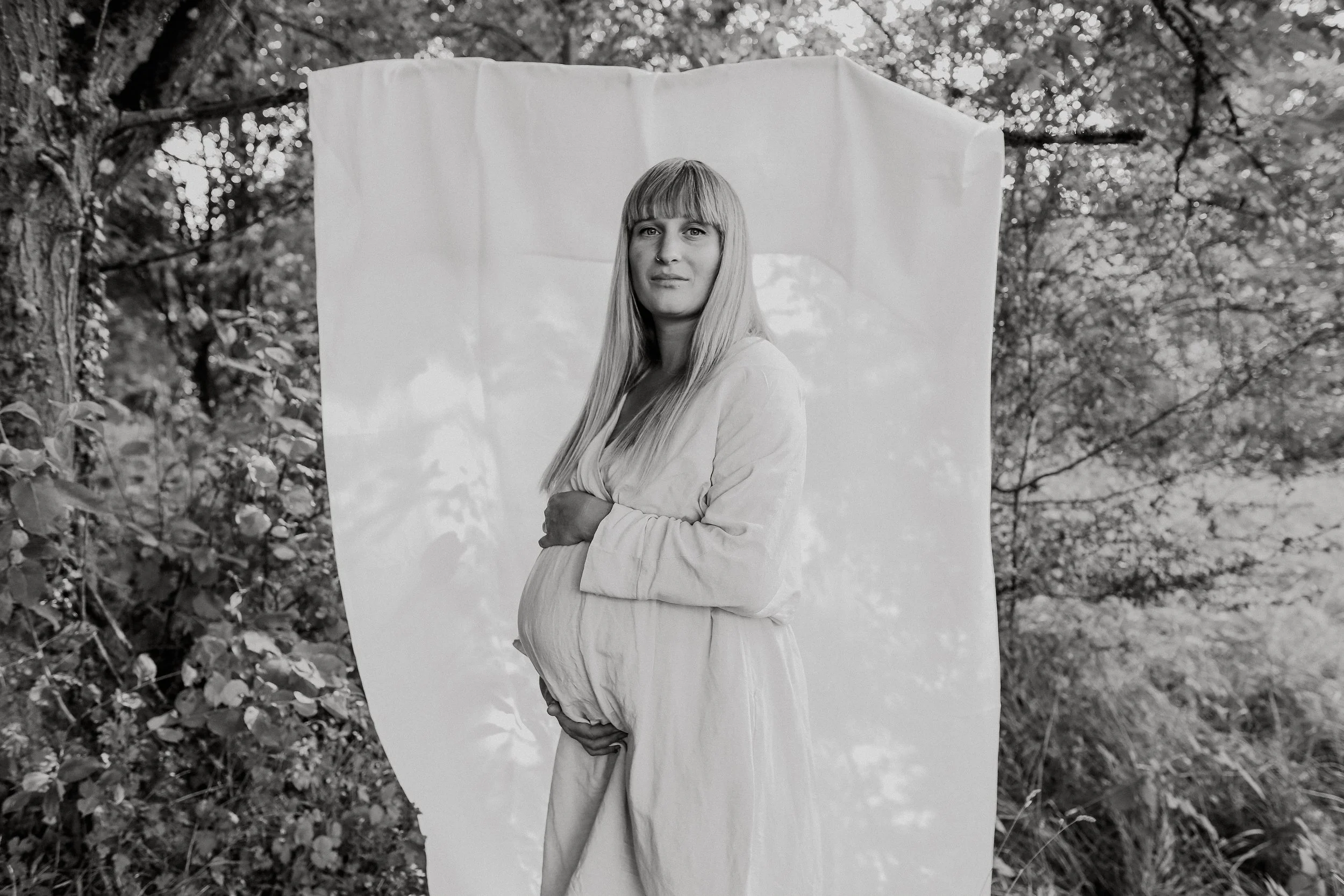 Schwangere Frau mit langen Haaren in einem weißen Kleid steht vor einem weißen Hintergrund im Freien, umgeben von Bäumen. Babybauchfotografie Saarland, Babybauchshooting Tageslichtstudio, Saarland, Schwangerschaftsfotografie, Babybauchfotografie, Fam