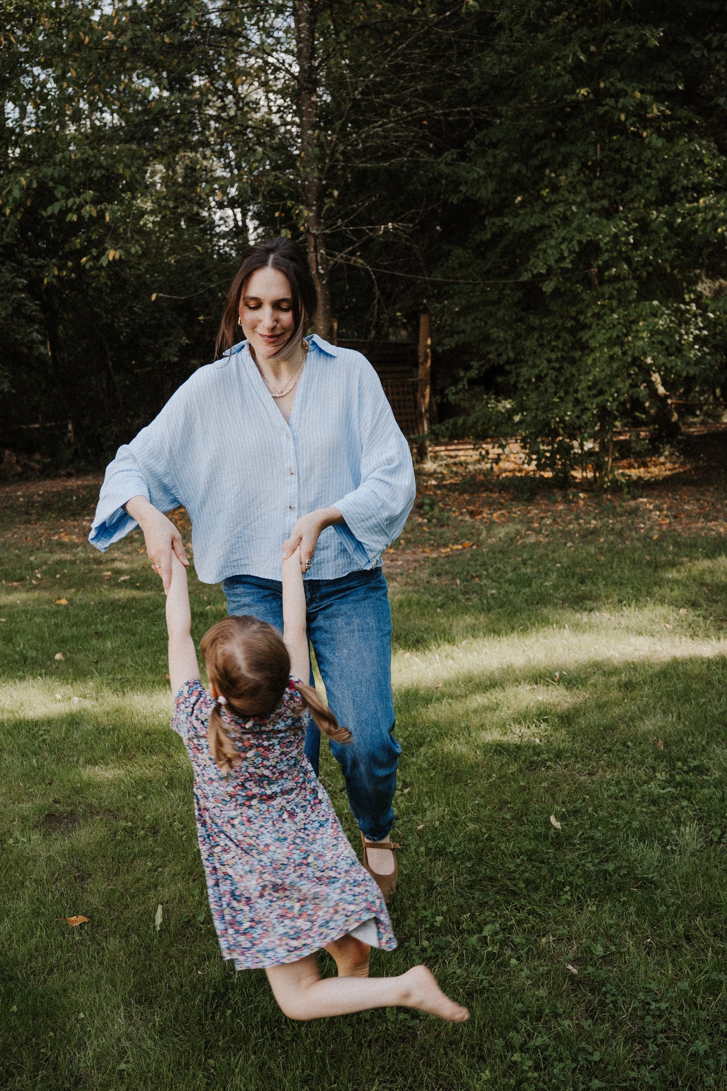 Eine Frau und ein Mädchen spielen im Garten, die Frau hält das Mädchen an den Händen, während das Mädchen fröhlich springt.Mutter & Kind Shooting im Saarland, Familienshooting im Saarland, Familienfotografin. Fotografie für Mütter und Kinder.