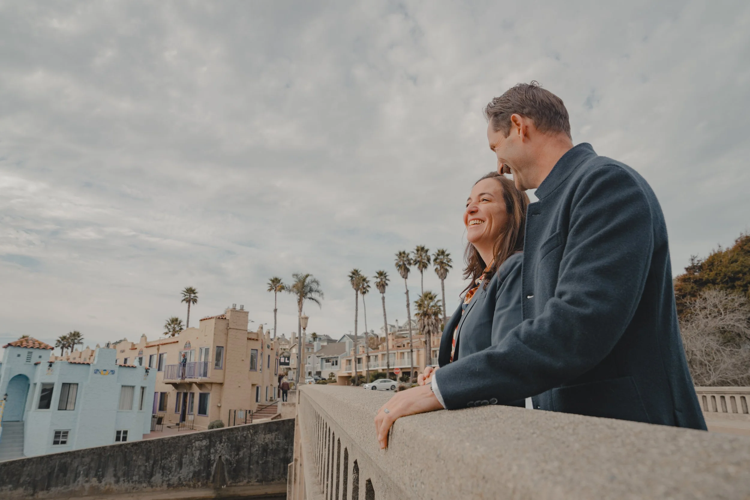 A man and a woman in Capitola Village on the bridge with buildings and trees in the background.