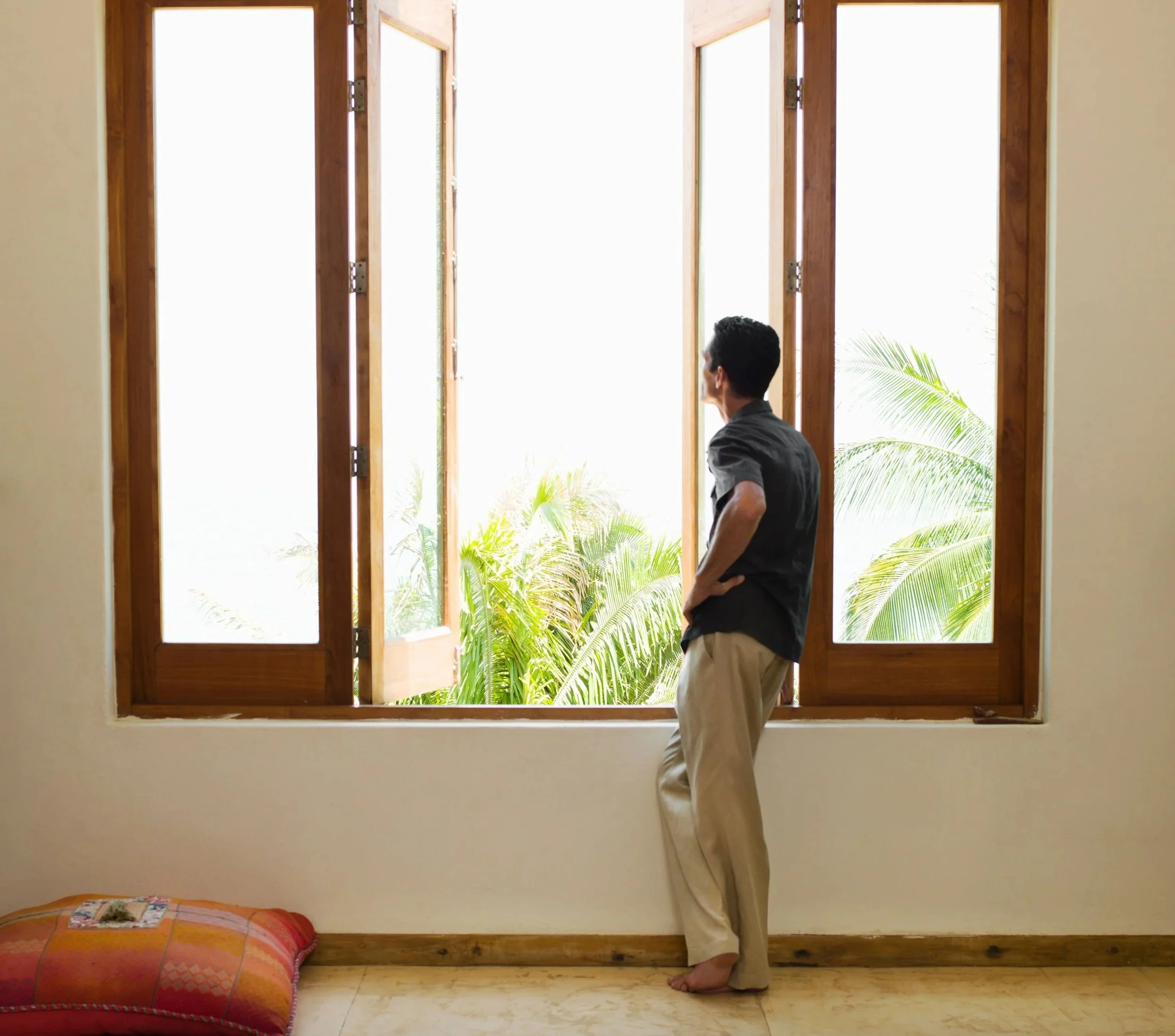 A man looking out a large window with palm trees.