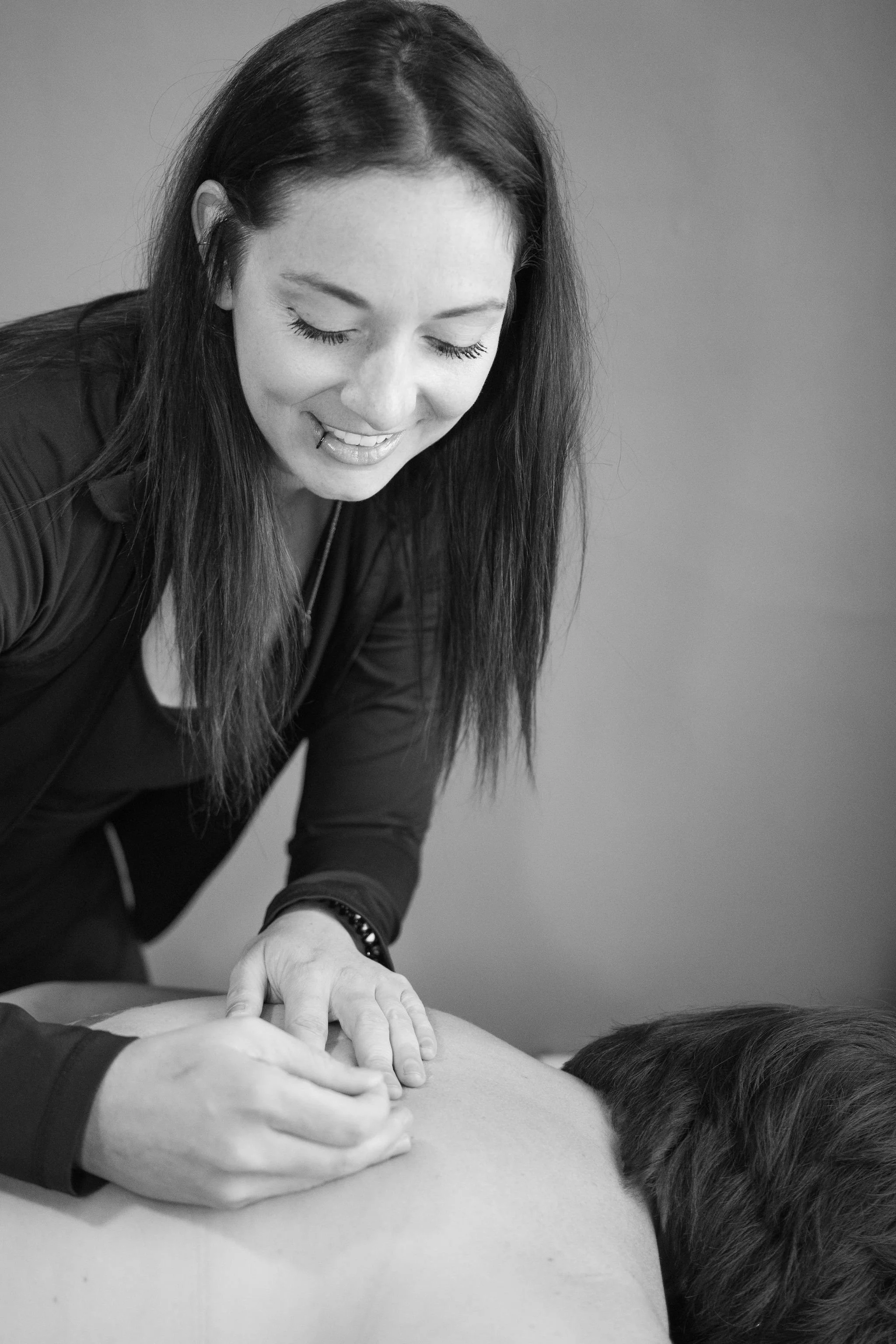 A woman with long dark hair giving a massage or physical therapy to a person's back, who is lying face down on a flat surface. The woman is smiling with her eyes closed.