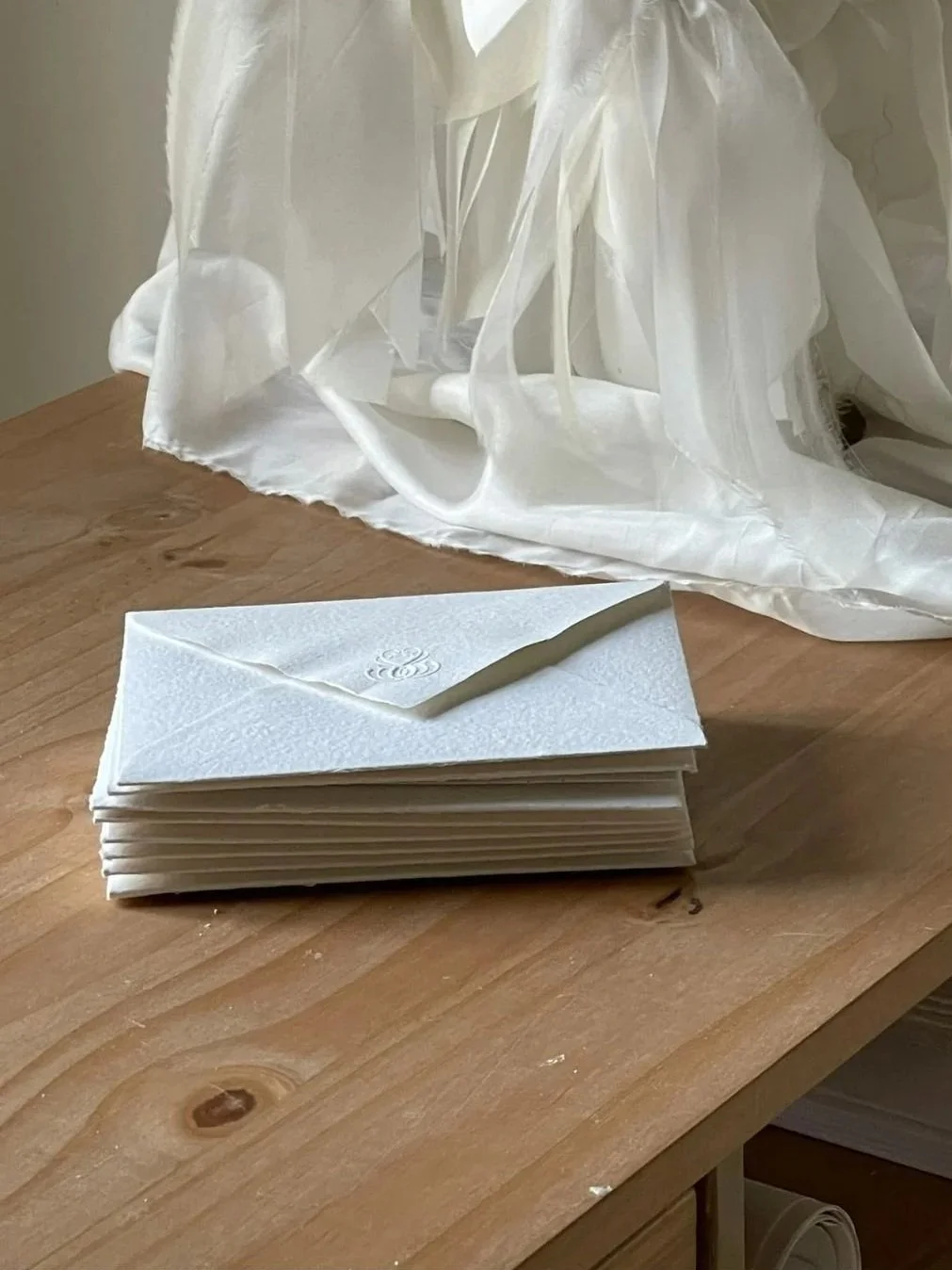 A stack of white envelopes and a pen on a wooden table, with a large, sheer white fabric draped in the background.