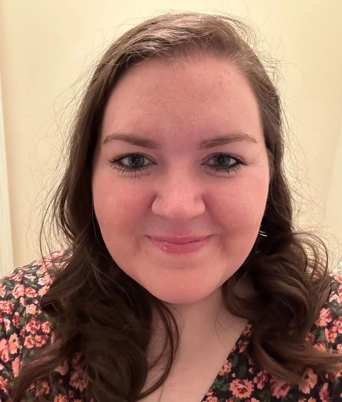 A woman with long, brown, curly hair smiling at the camera. She has blue eyes and is wearing a floral patterned top.