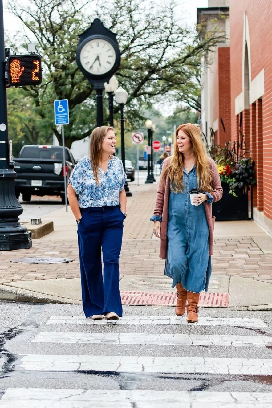 Two women walking and talking on a crosswalk in an urban area during daytime, with one holding a coffee cup, seen behind a clock and traffic signals, with trees and brick buildings nearby.