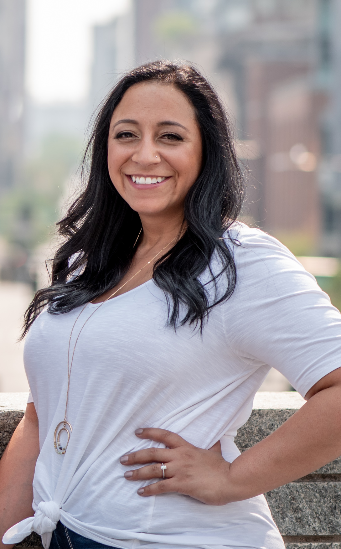 Portrait of a woman with long dark hair, wearing a white knotted t-shirt, standing outdoors with buildings blurred in the background.