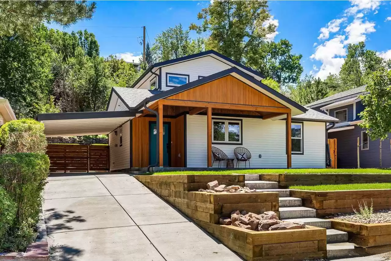 Modern house with white siding, wooden accents, a covered porch with outdoor chairs, a sloped driveway, landscaped yard with rocks and plants, and surrounded by trees.