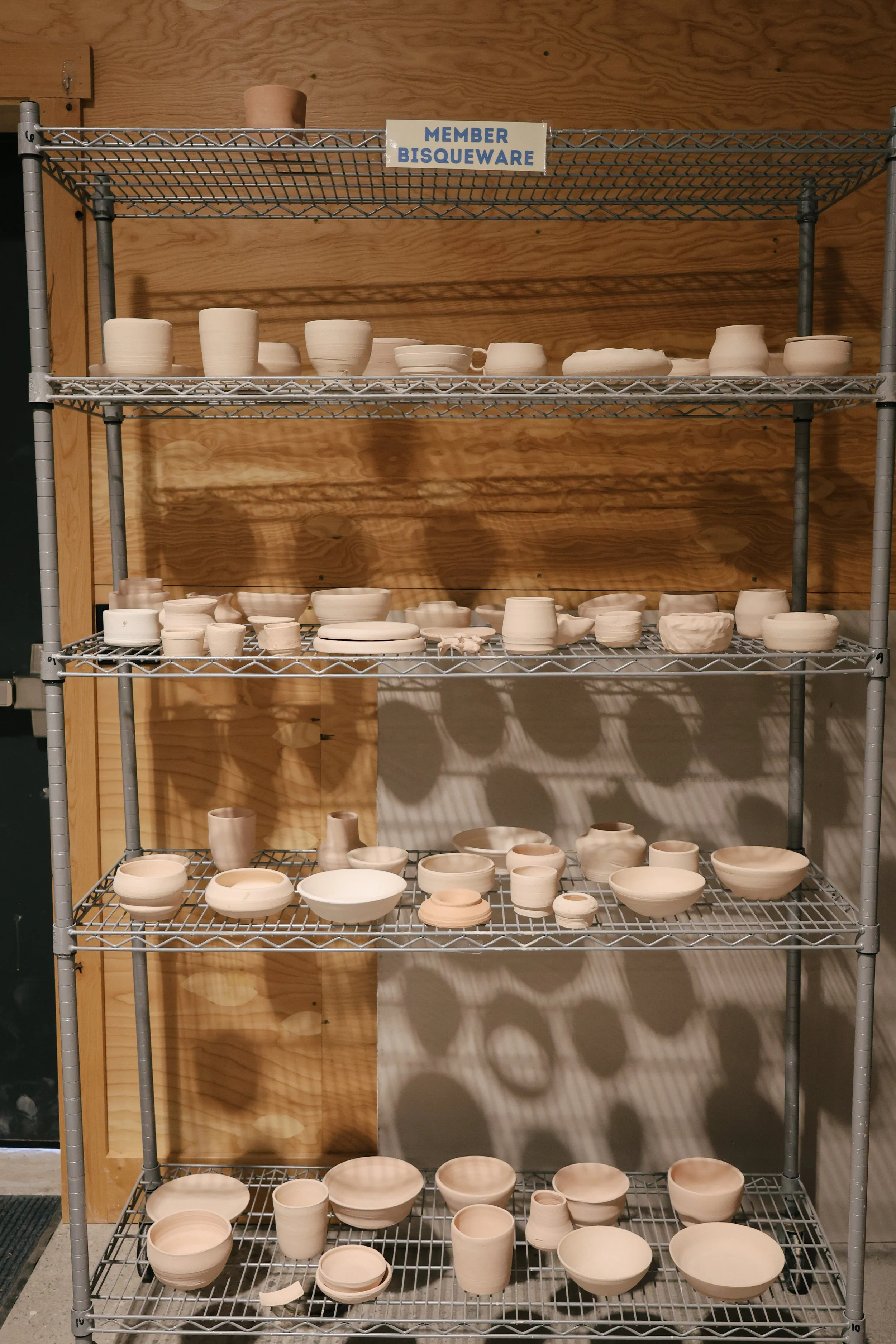 Metal shelving unit with four shelves holding unglazed ceramic bowls, cups, and plates in various shapes and sizes, set against a wooden wall. A sign on the top shelf reads 'Member Bisqueware'.
