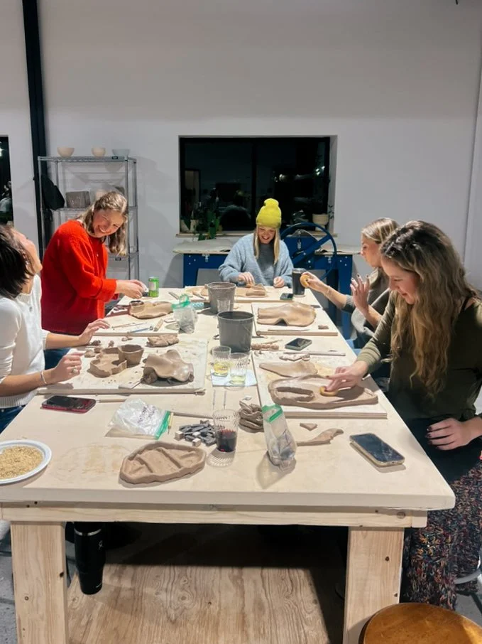 Group of women gathered around a table, working on pottery-making projects in a studio, with clay and tools on the table.