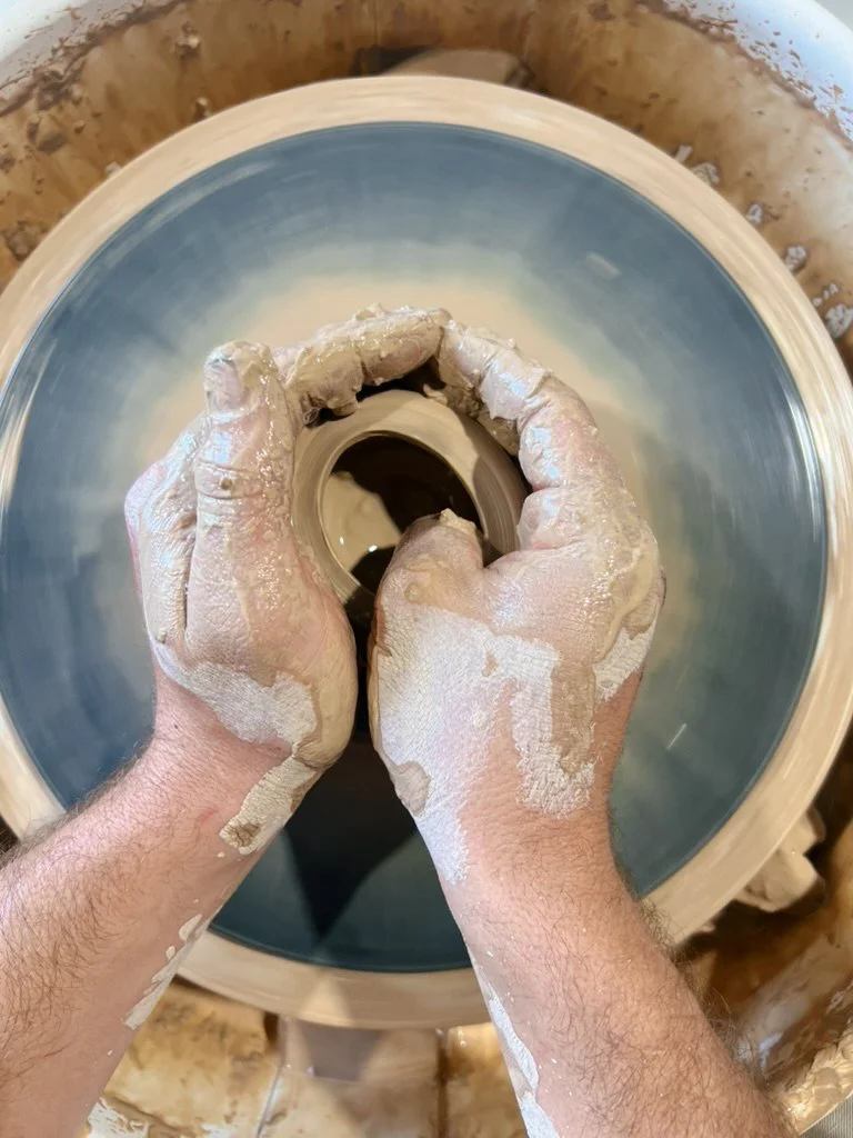 Photograph of a person shaping clay on a pottery wheel, with hands covered in wet clay.