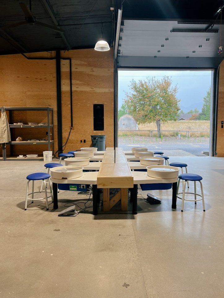 An indoor pottery studio with a long wooden table set up for pottery wheel work, surrounded by blue-cushioned stools, with a view of an open garage door showing an outdoor scene with a tree and a building.
