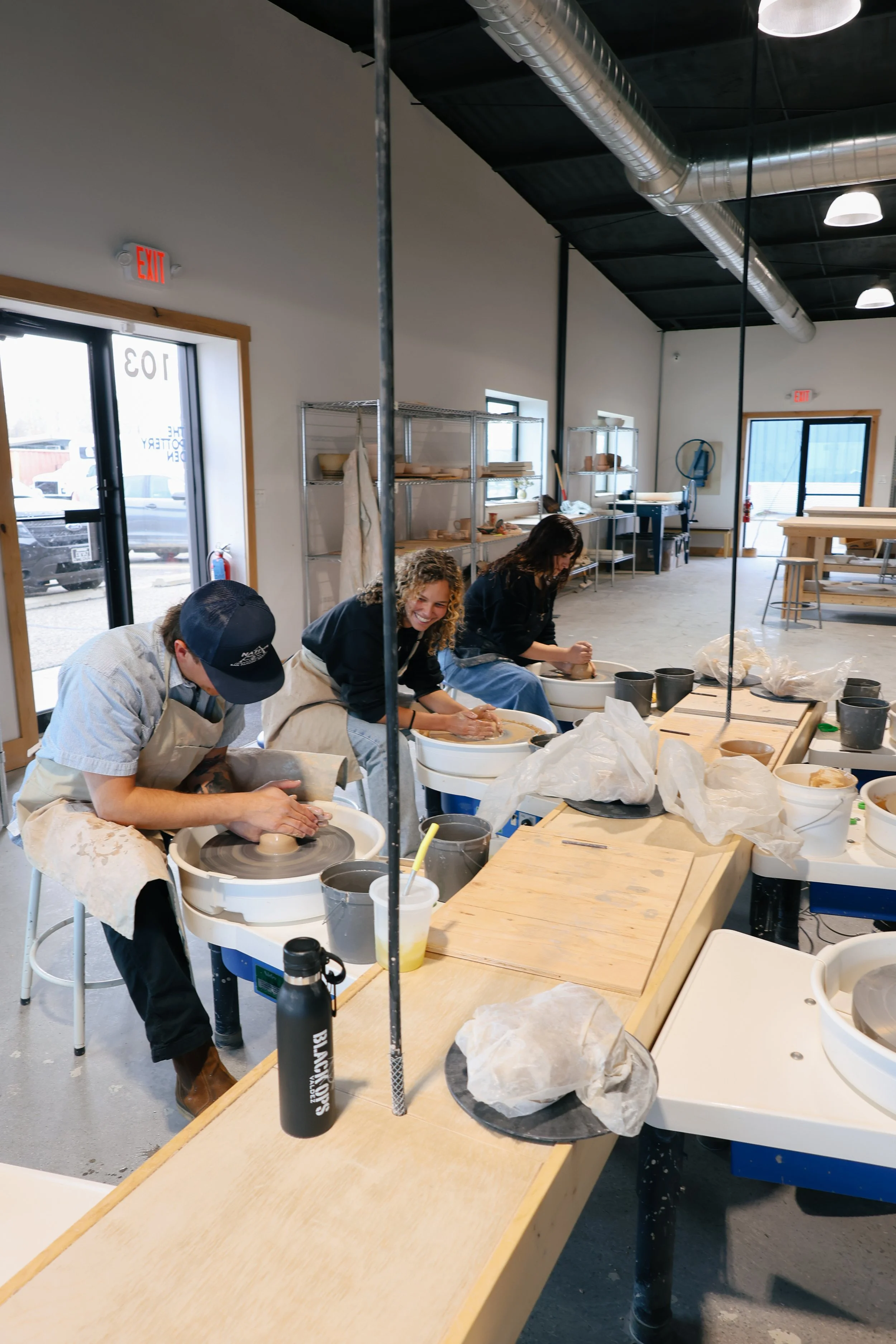 People working at pottery wheels in a ceramics studio with shelves of clay and supplies, large windows, and metal ventilation ducts.