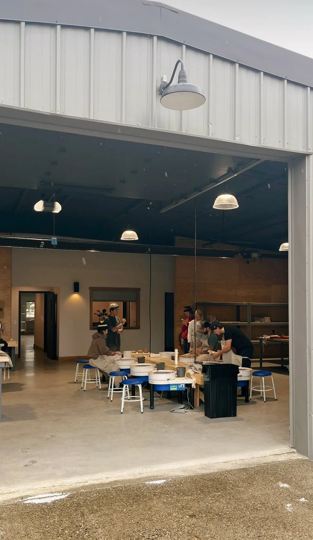 Inside a workshop or classroom, several people are working at a long table with pottery wheels and clay. Large mirrors reflect the scene, and the area is lit by hanging industrial lights. The space has wooden and concrete walls, and an open garage-style door reveals an outdoor area with wet pavement.