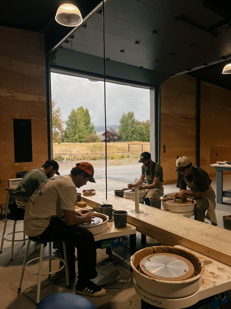 Four people engaged in pottery making at a ceramic studio, sitting around a wooden table, with large windows showing a scenic outdoor landscape.