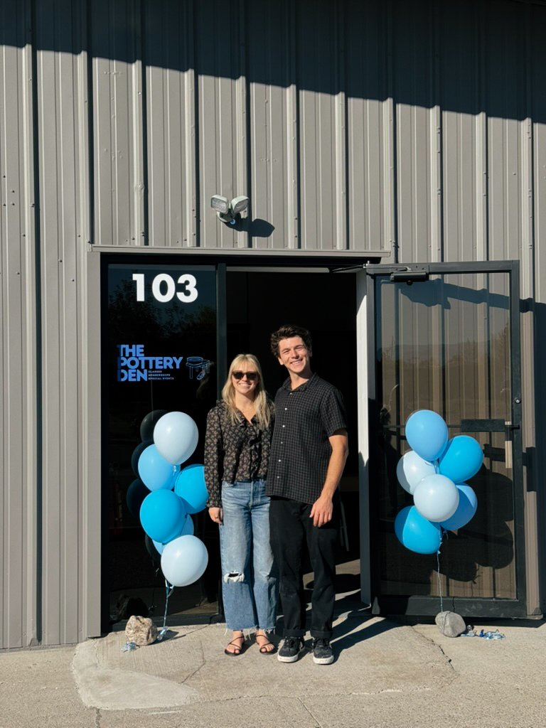 Two people standing outside a building entrance decorated with blue and white balloons for a celebration, with the sign 'The Pottery Den' visible on the glass door.