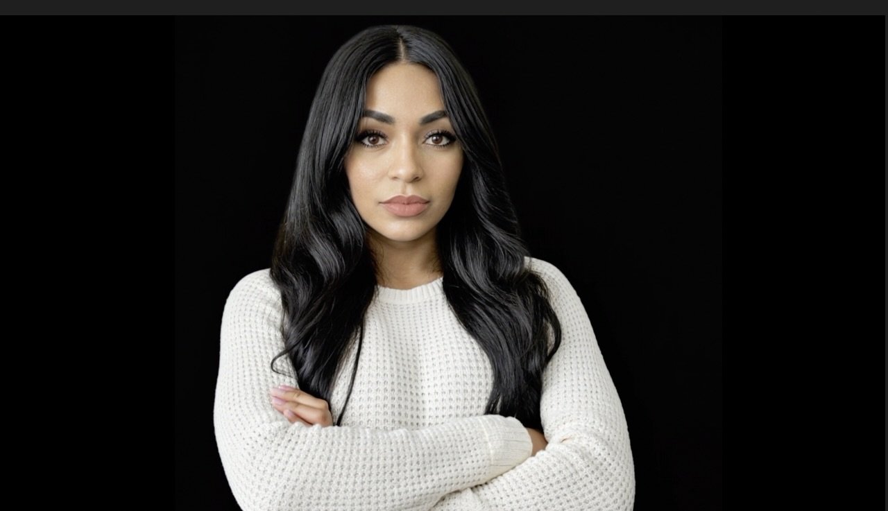 Close-up of a woman with long black hair, wearing a white knit sweater, arms crossed, against a black background.