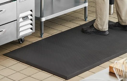 Part of a person standing on a black rubber mat on a brown tiled floor in a kitchen or food service area.