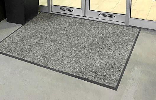 A gray rectangular doormat placed in front of glass door entrance, with a black border and a small black planter in the corner.