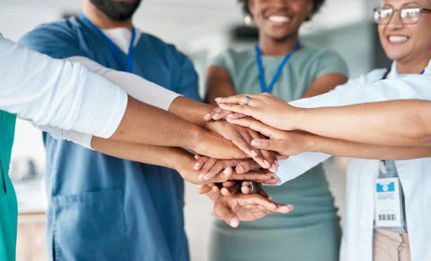 Healthcare workers and staff from different backgrounds placing their hands together in a gesture of teamwork.