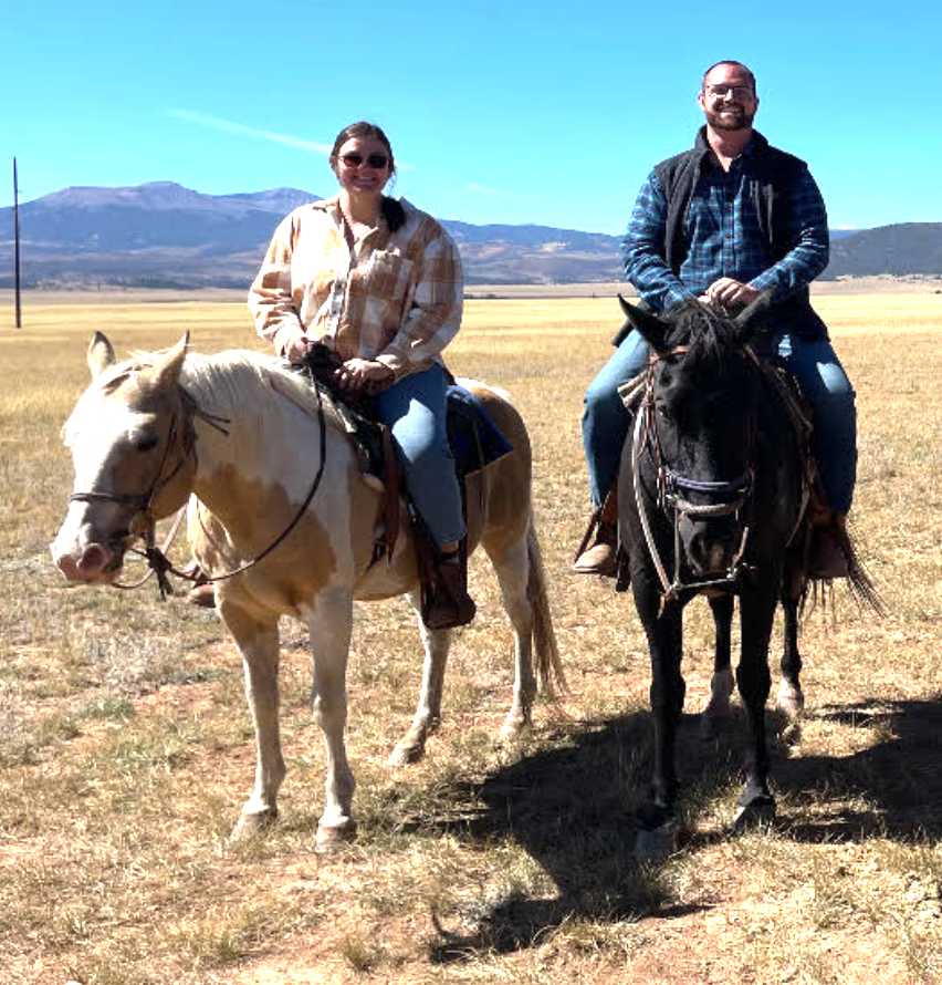 A man and woman riding horses in an open field with mountains in the background.