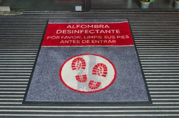 Doormat with a red and white sign reading 'Alfombra Desinfectante Por Favor, Limpie Sus Pies Antes de Entrar' and footprints in a circle, placed on a striped floor.