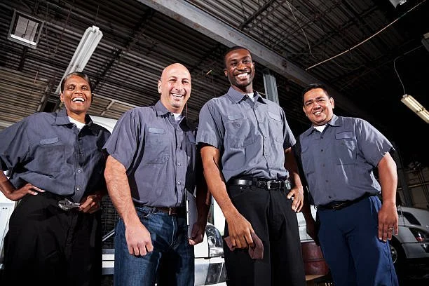 Four male automotive technicians in uniform standing in a car repair garage, smiling at the camera.