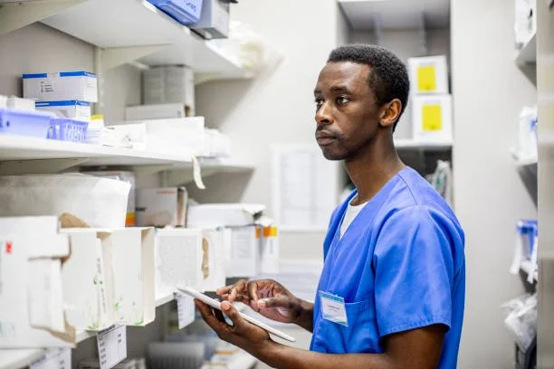 Male healthcare professional in blue scrubs looking at medication shelves in a pharmacy or hospital storage room while holding a clipboard.