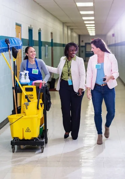 Three women walking down a hospital hallway, one with a cleaning cart, all smiling and engaged in conversation.