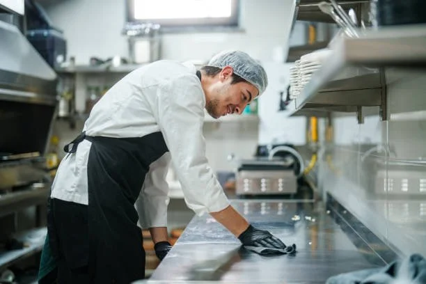 A chef working in a commercial kitchen, cleaning the stainless steel counter.