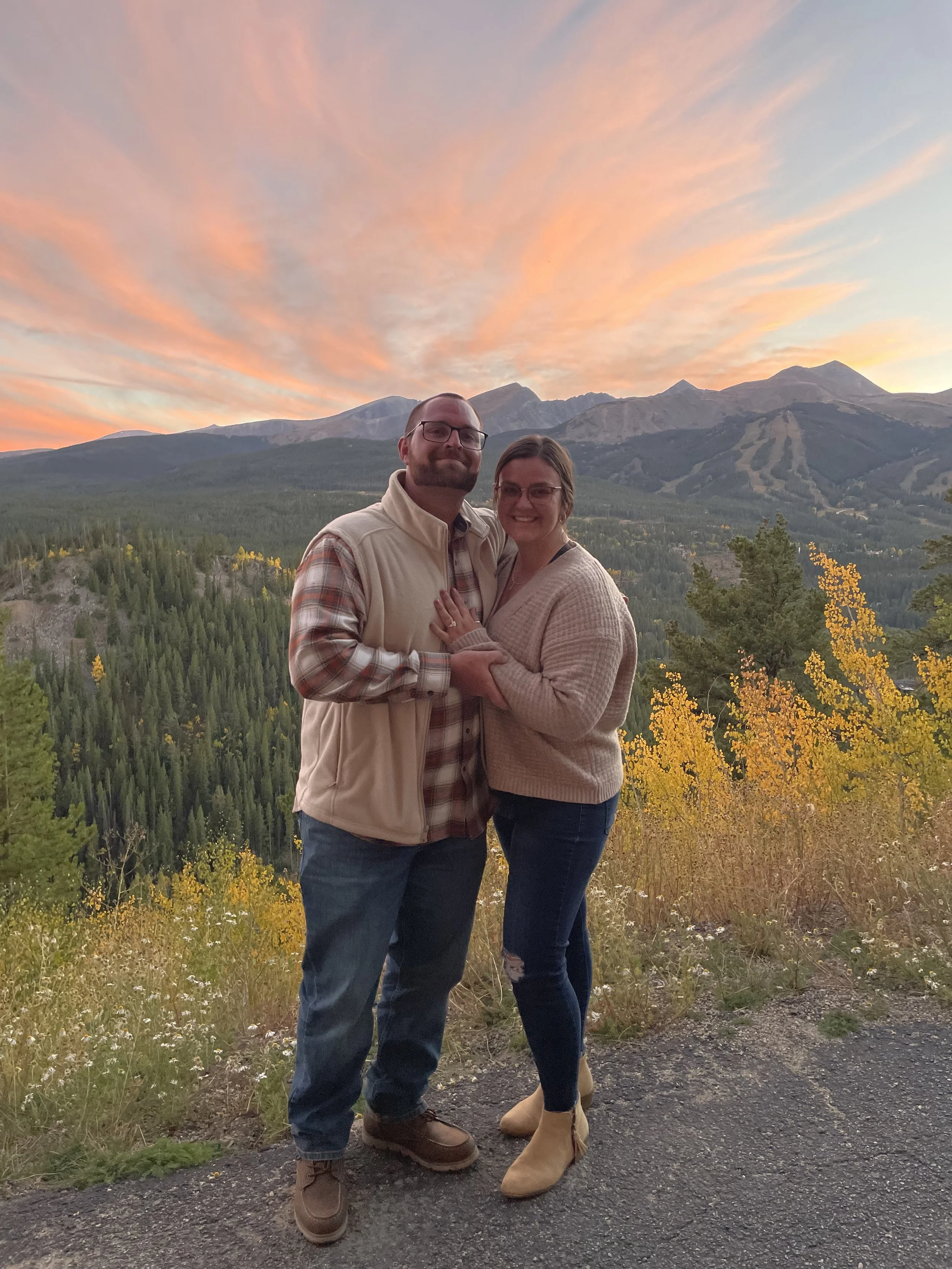 A couple standing close together outdoors during sunset, with mountains and a colorful sky in the background.