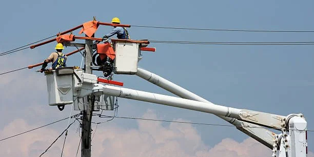 Two utility workers wearing yellow helmets repairing power lines from a bucket lift against a cloudy sky.