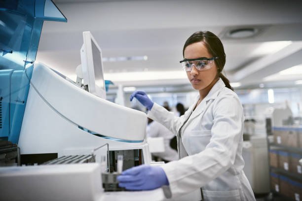A female scientist in a white lab coat, safety goggles, and blue gloves working with laboratory equipment in a modern laboratory setting.