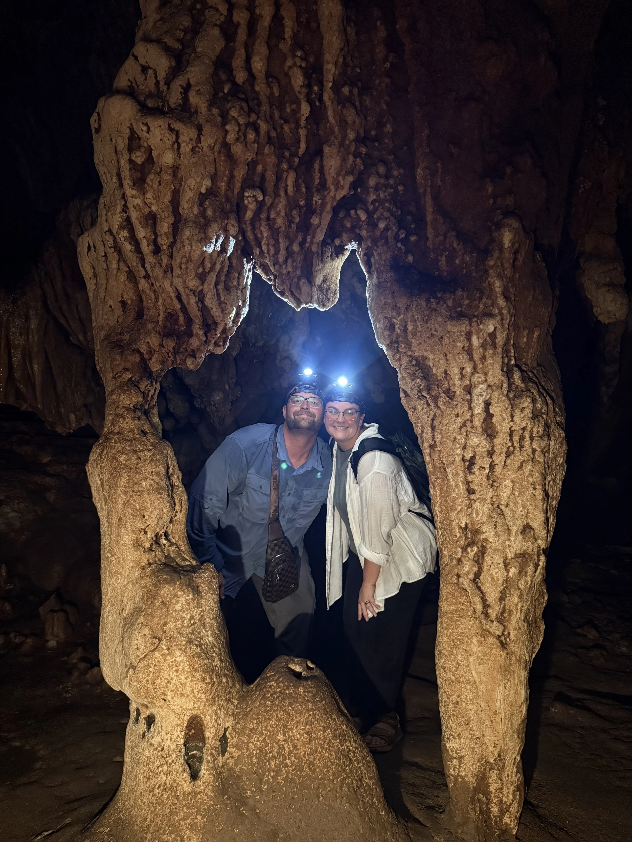 Two people, a man and a woman, are inside a cave and smiling at the camera. They are wearing headlamps, and the cave has stalactites and stalagmites with a rough, textured surface.