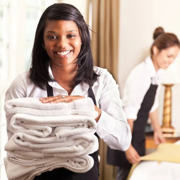 A smiling woman holding a stack of folded white towels in a restaurant or hotel setting, with another woman in the background preparing a table.