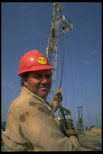 A worker wearing a red safety helmet and beige work uniform standing outdoors near a tall metal communication tower against a blue sky.