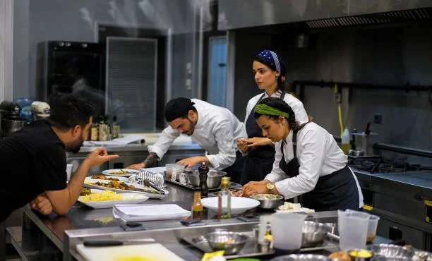 Four chefs preparing food in a commercial kitchen, two women and two men, with various cooking utensils and ingredients on the counter.