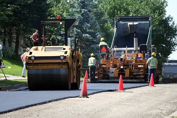 Construction workers paving a road with asphalt using a roller and paver, with orange cones marking the area.