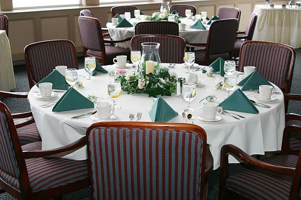 A round table set for a formal event with a white tablecloth, turquoise napkins, white cups and saucers, glasses of water, and a floral centerpiece with candles. Multiple chairs surround the table in a room with large windows and additional tables in the background.