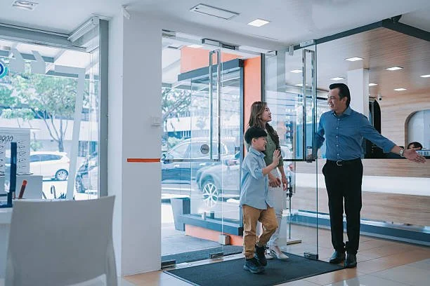 A man and two children entering a modern building through glass doors, with the man greeting them inside.