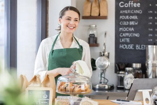 Smiling woman wearing a green apron behind the counter of a coffee shop, with a display case of baked goods and a menu board in the background.