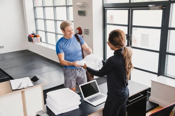 A woman behind a reception desk handing a paper package to a smiling man in a blue shirt at a modern office or gym reception area.