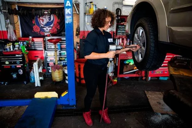 A woman changing a tire on a vehicle in an auto repair shop. She is holding a tool and working on the car's wheel while standing on the shop floor, surrounded by tools and equipment.