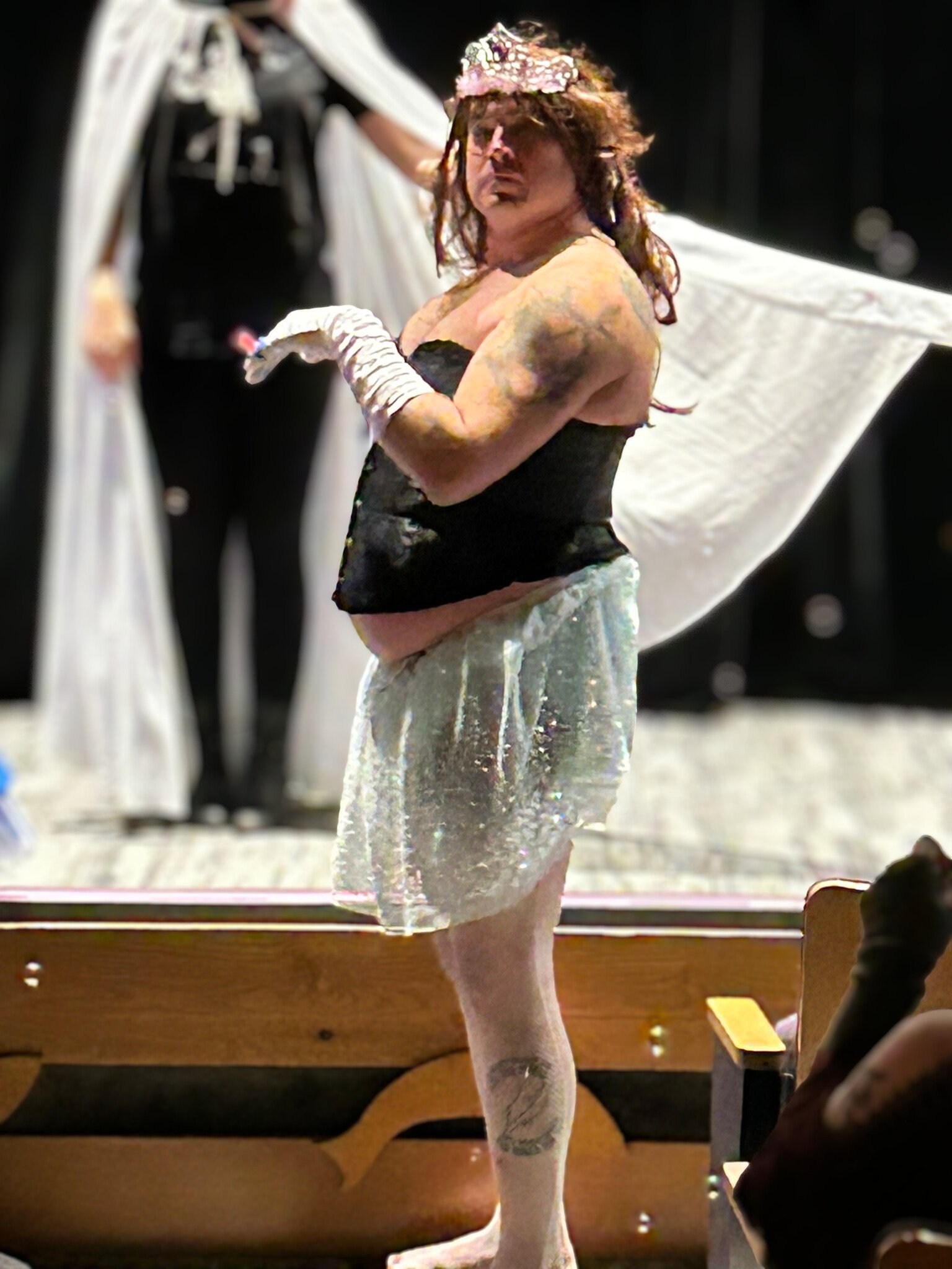 A performer dressed in a corset, sparkly skirt, long white gloves and a tiara stares seriously into the camera while behind them on-stage is another performer extending a sheer white cape.