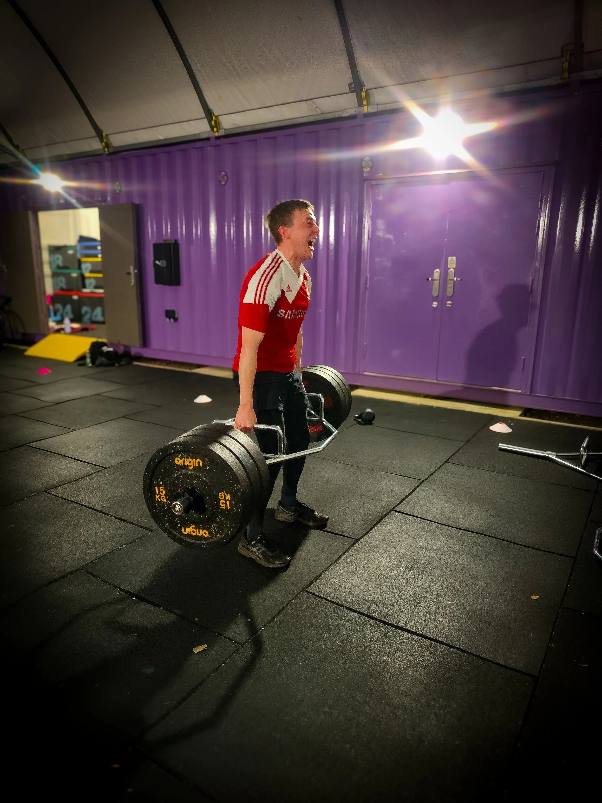 A man lifting a heavy barbell with black weights in a gym, smiling and wearing a red athletic shirt and black pants.