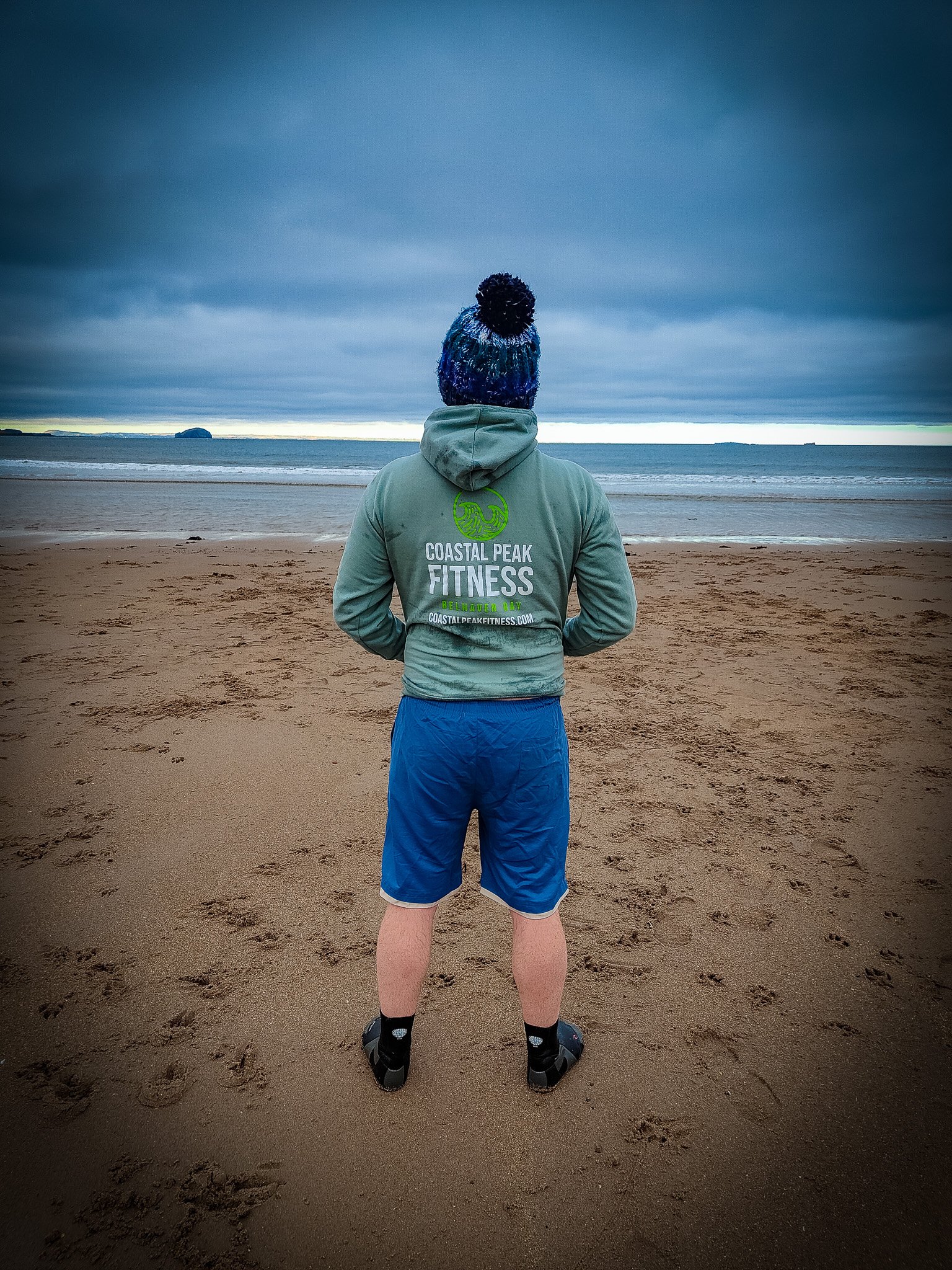 A person standing on a beach facing the ocean, wearing a green hoodie, blue shorts, black sandals, and a multicolored knit beanie with a pom-pom, under a cloudy sky.