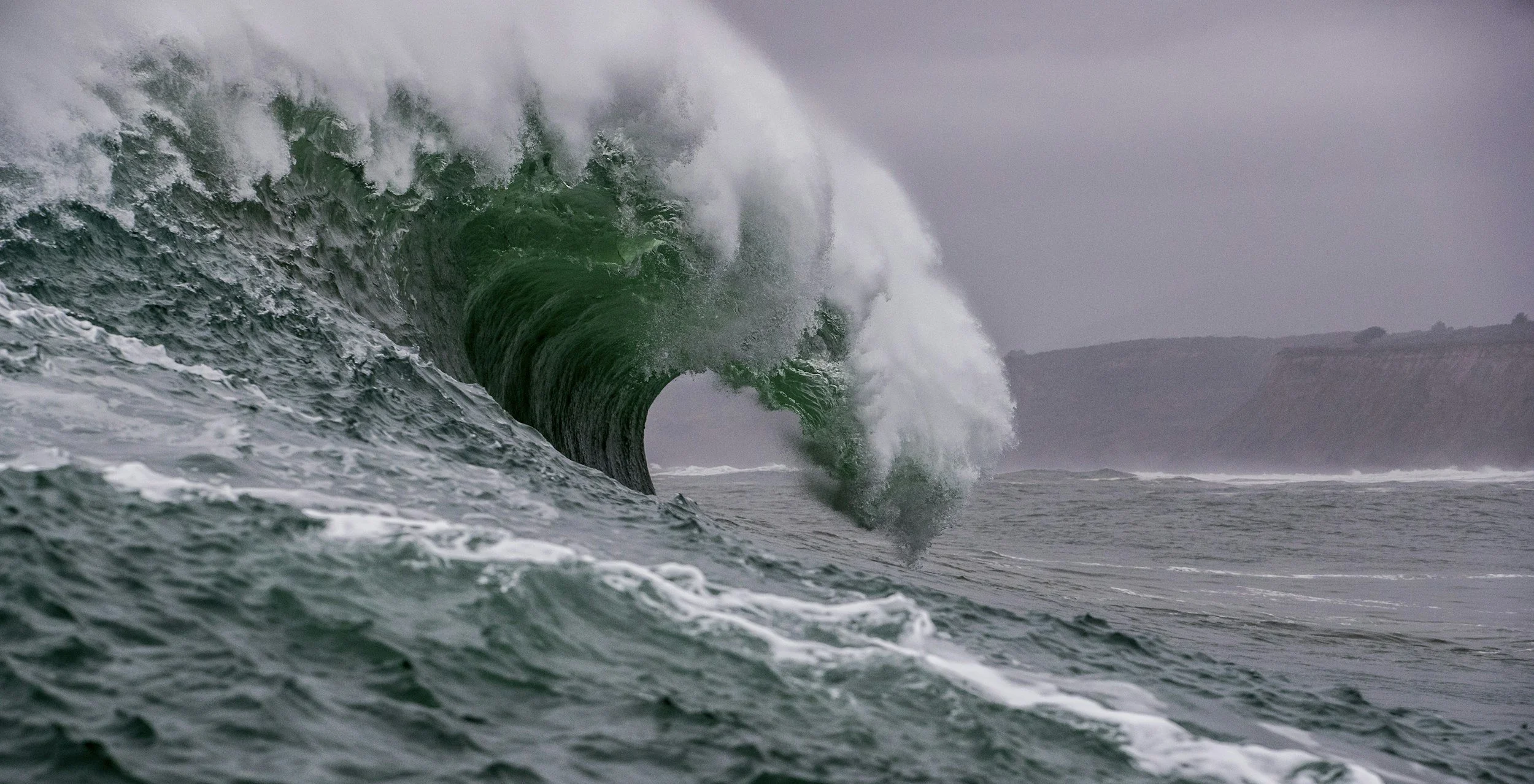 Large ocean wave approaching the shore with a cliff in the background under a cloudy sky.