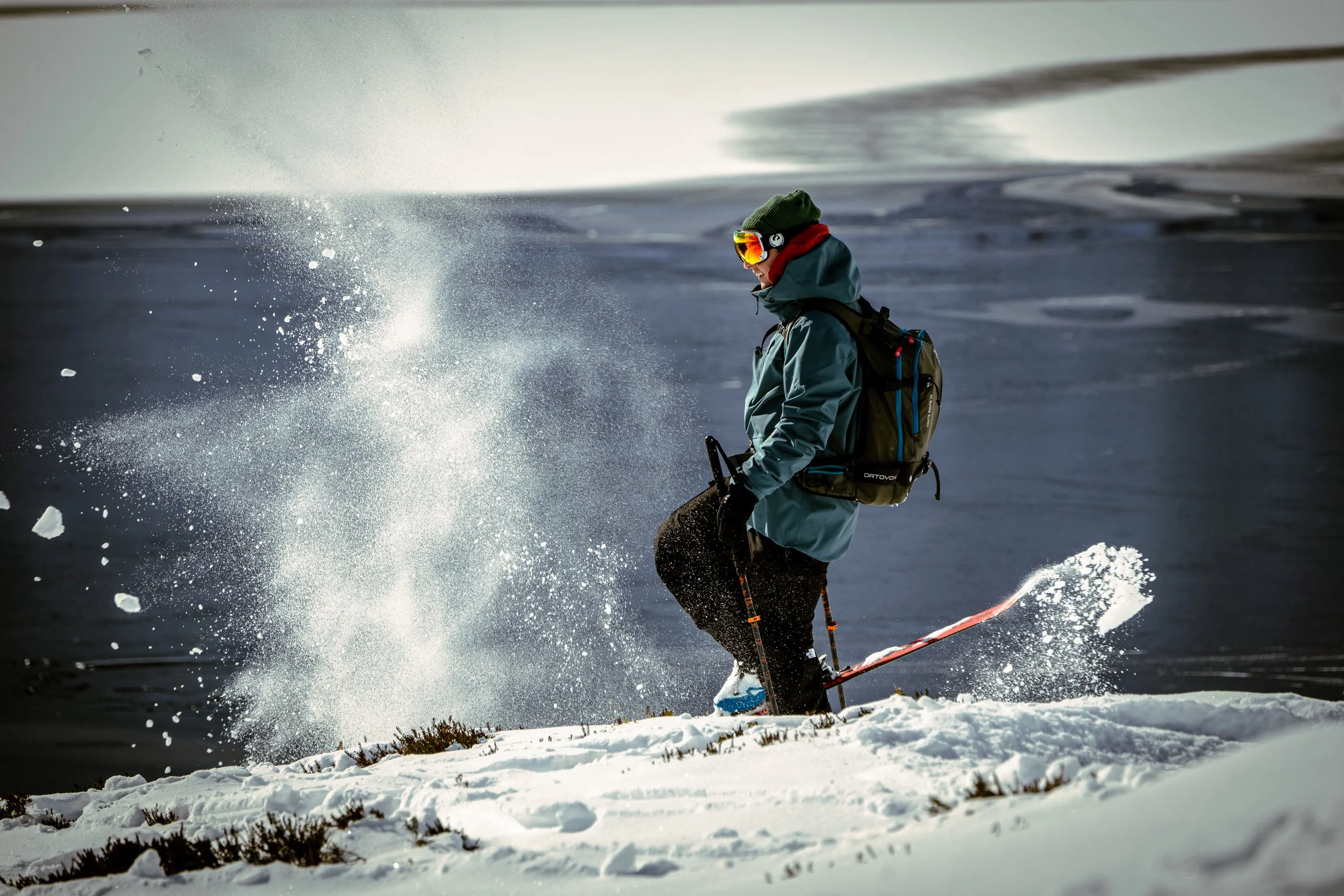 Person snowboarding on snow-covered terrain near water, wearing a jacket, hat, goggles, and carrying a backpack.