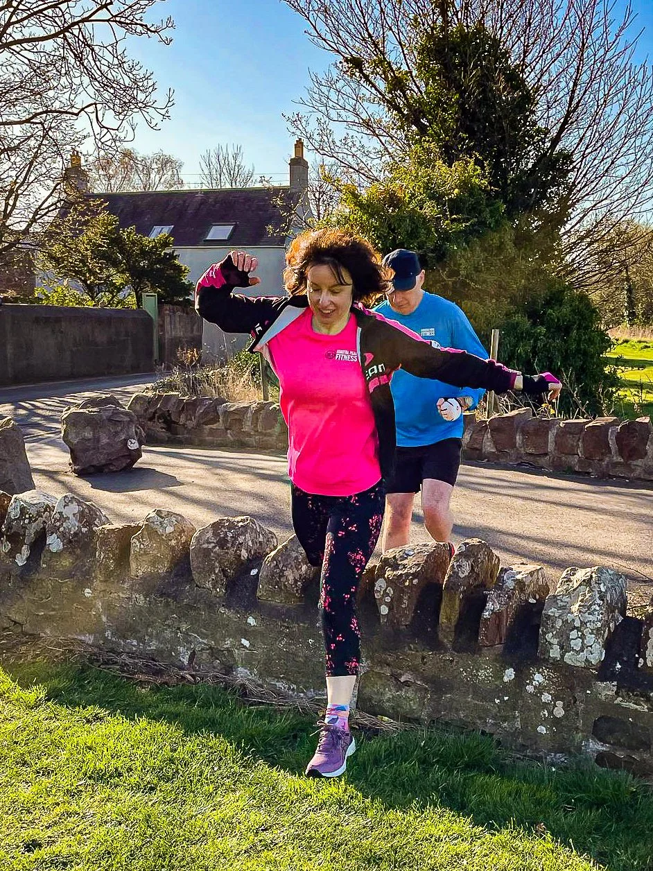 Female runner in pink top jumping over a stone wall in the sun