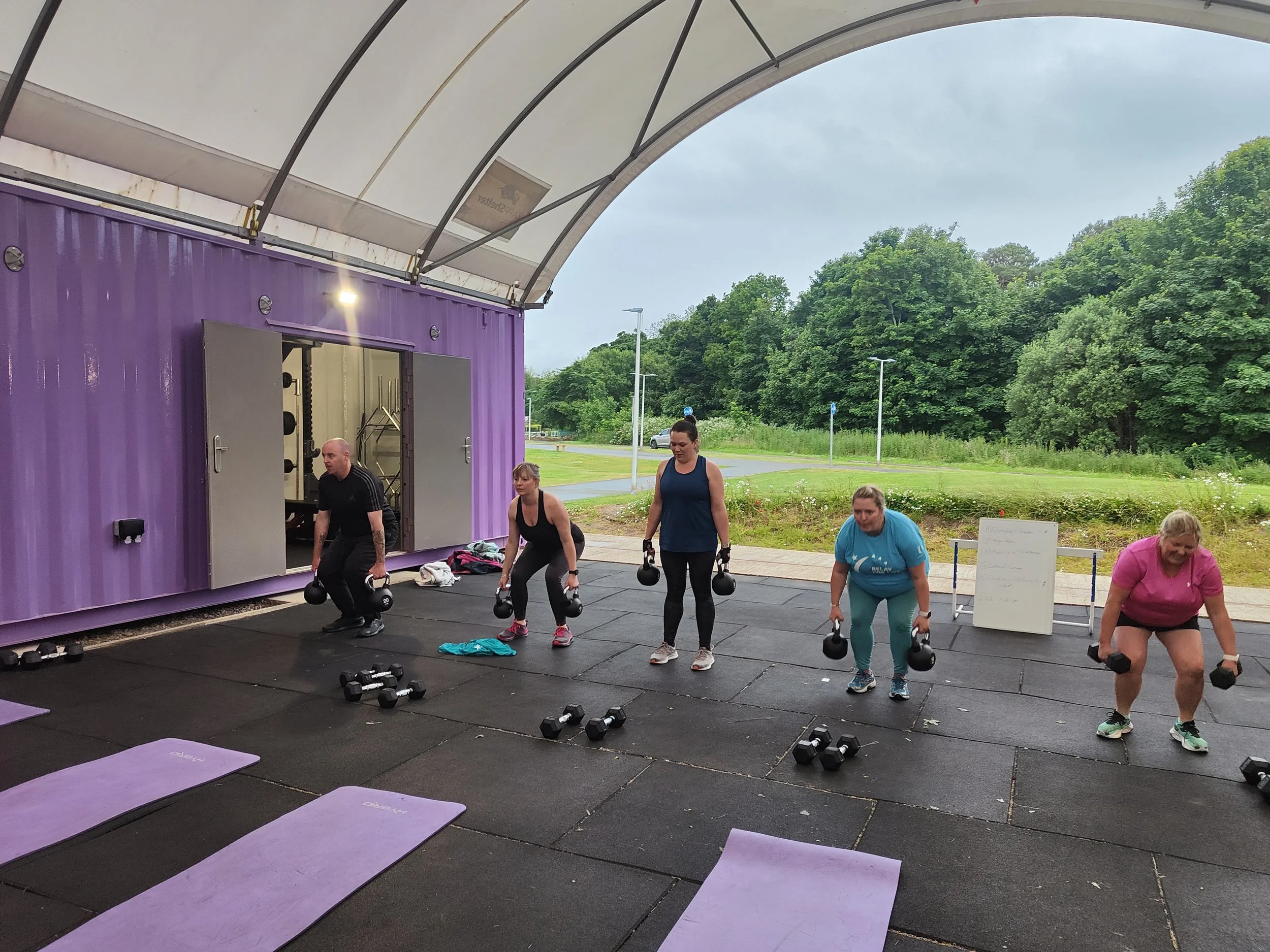 People participating in an outdoor workout class, lifting dumbbells on a rubber mat surface under a covered area with green trees in the background.